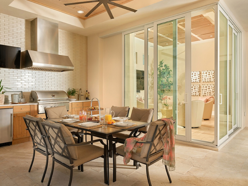 Bright modern dining area with a glass table, metal chairs, and sunlit kitchen opening to an enclosed patio.