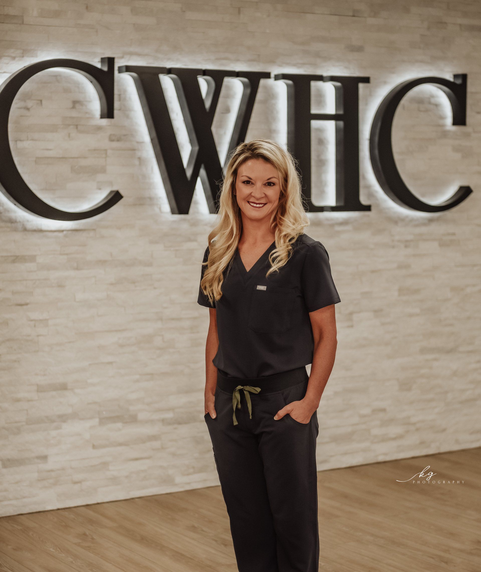 A professional standing in dark scrubs with hands in pockets before a wall featuring the lit, black sign 