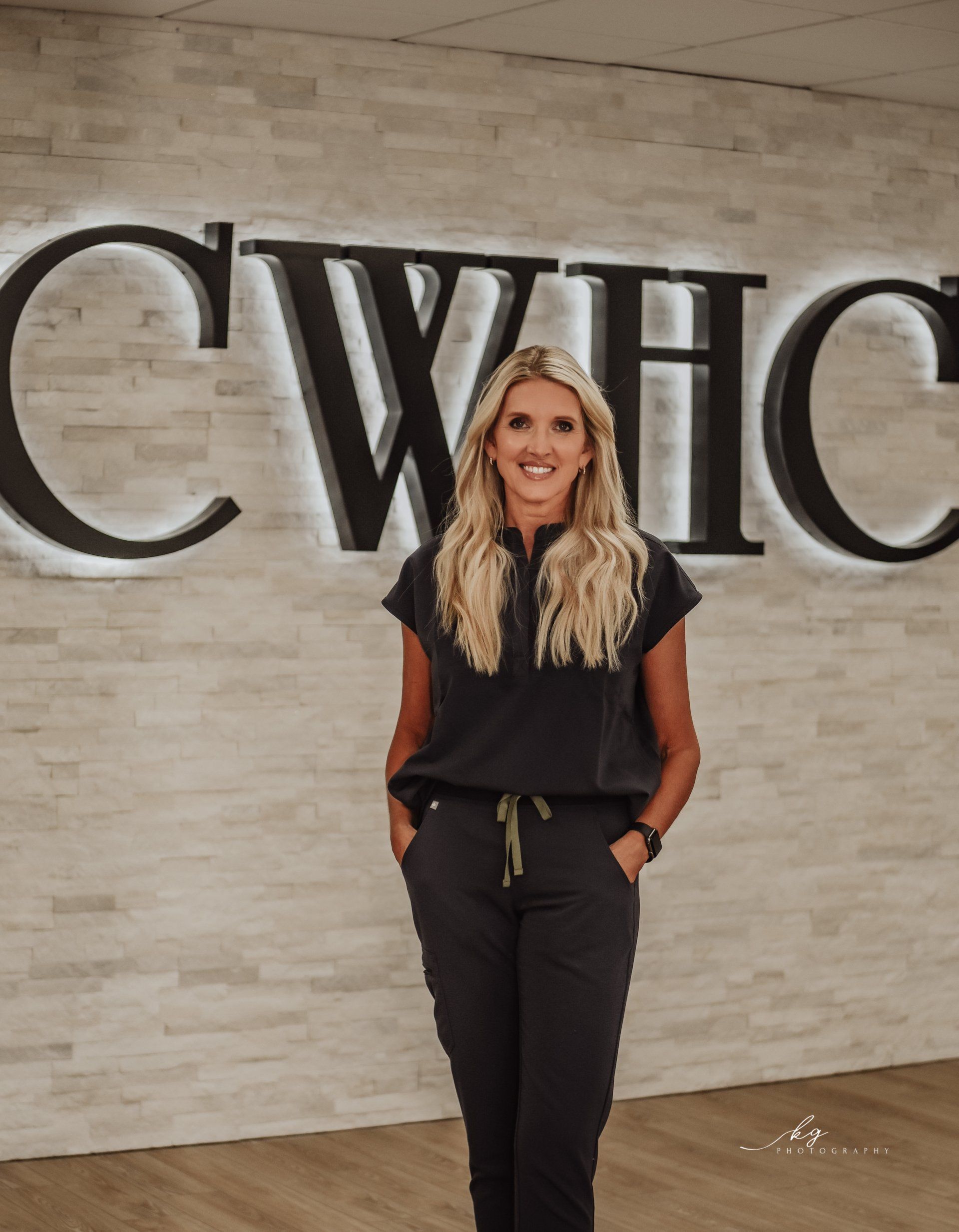 A person smiling in dark scrubs, standing in front of a white stone wall with the black letters CWHC illuminated.