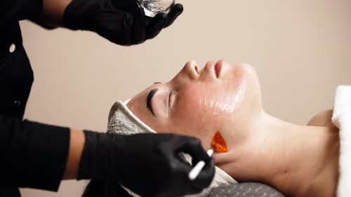 An esthetician in black gloves uses an orange applicator brush to apply a facial treatment to a client’s skin.