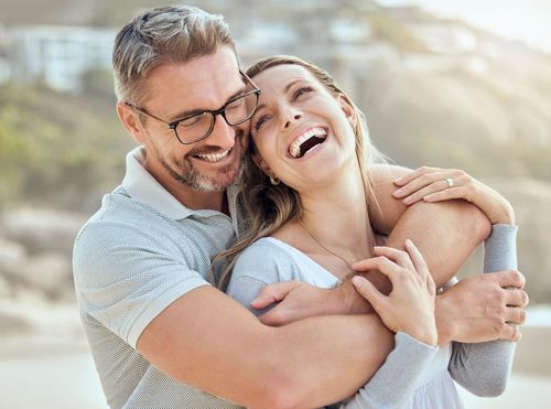 A person wearing glasses smiles while embracing another person who is laughing during an outdoor beach scene.