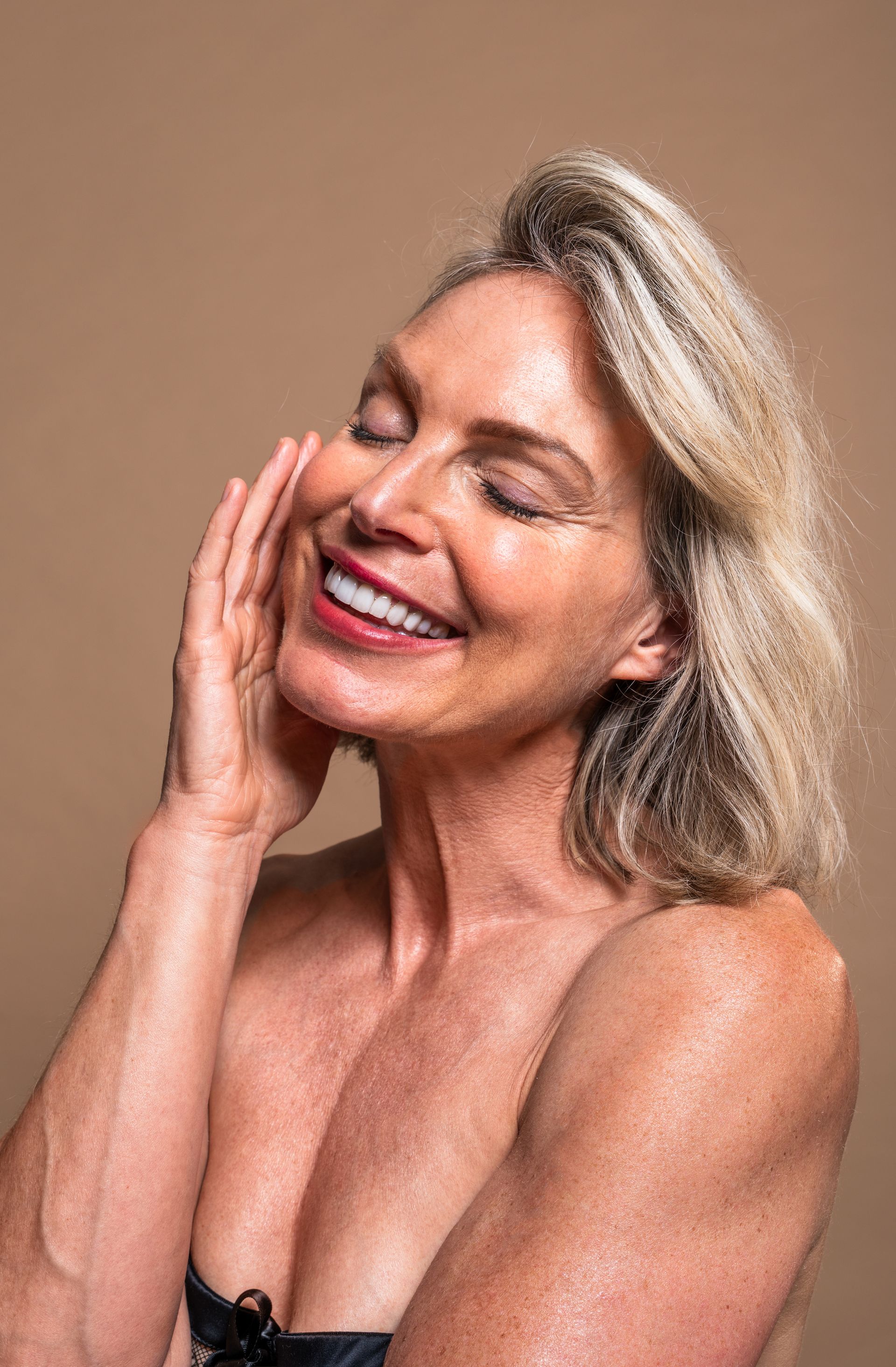 A person with blonde hair and a radiant expression touches their cheek against a warm-toned, solid background.