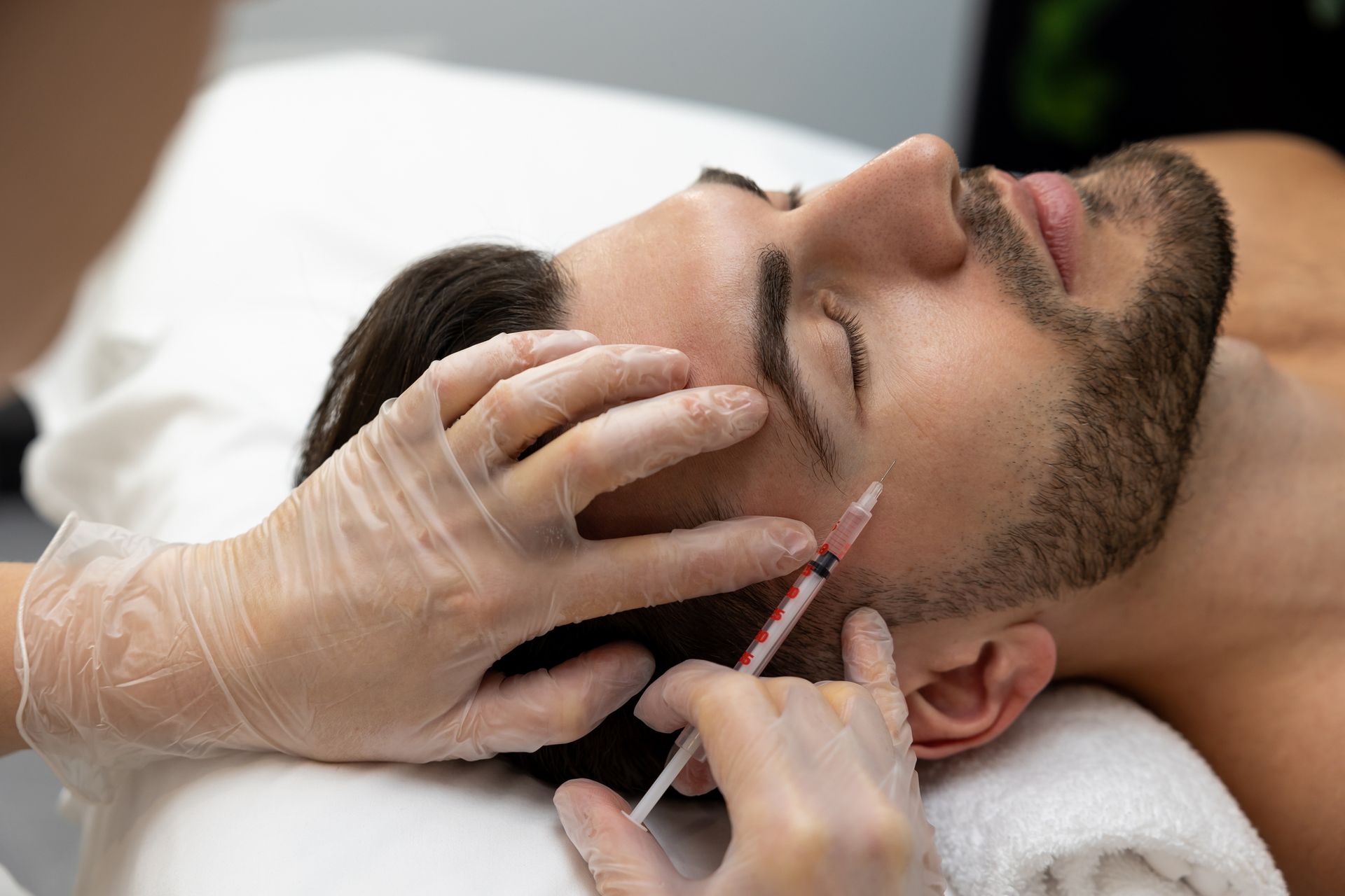 A provider in gloves injects a cosmetic treatment into a person's temple area while the person lies on a treatment table.