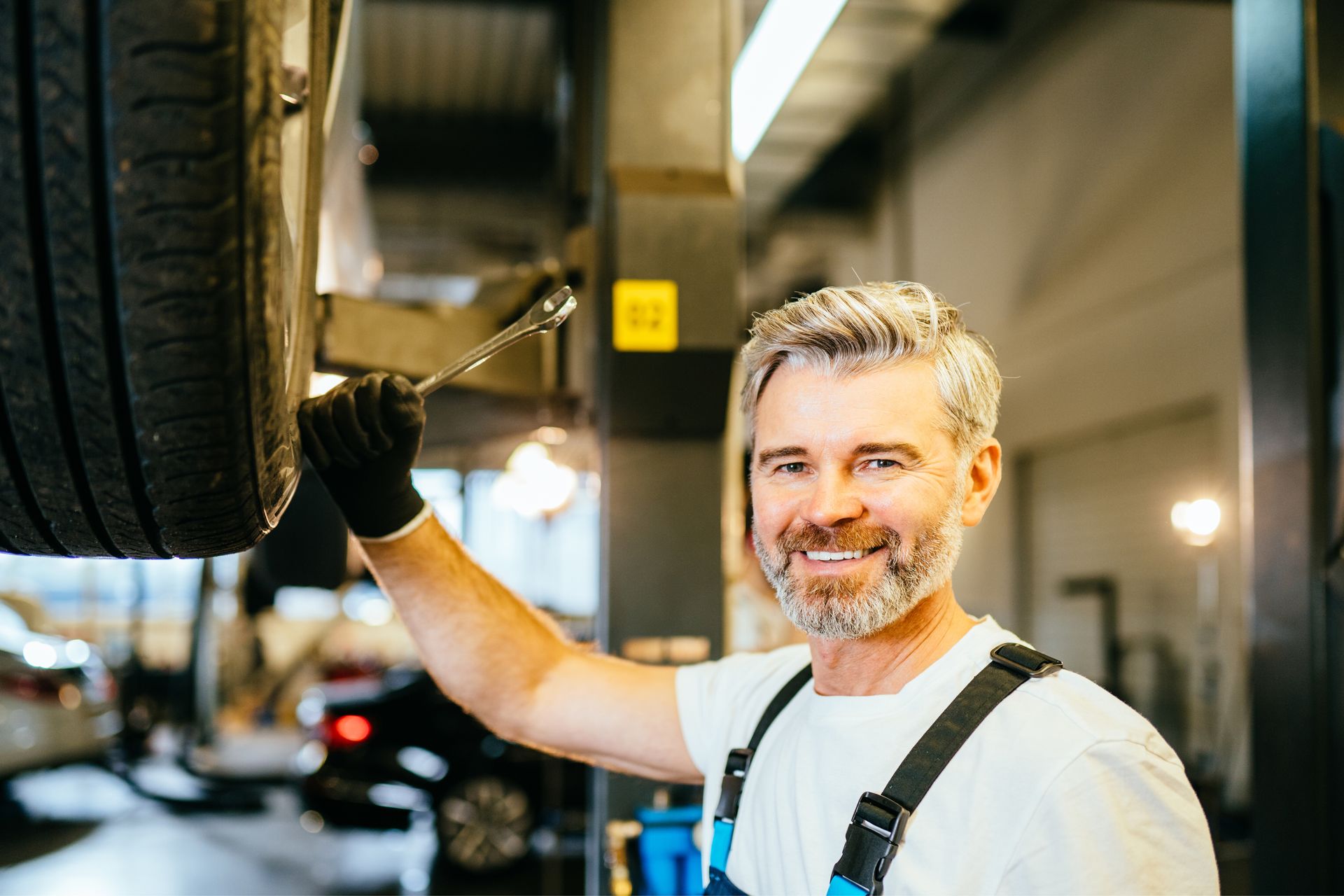 Mechanic with gray hair and a beard, wearing a white shirt and blue overalls, smiles while holding a tool by a car tire.