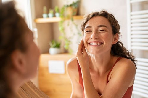 A smiling person with curly hair looks at their reflection in a mirror, touching their cheek in a bright, indoor setting.
