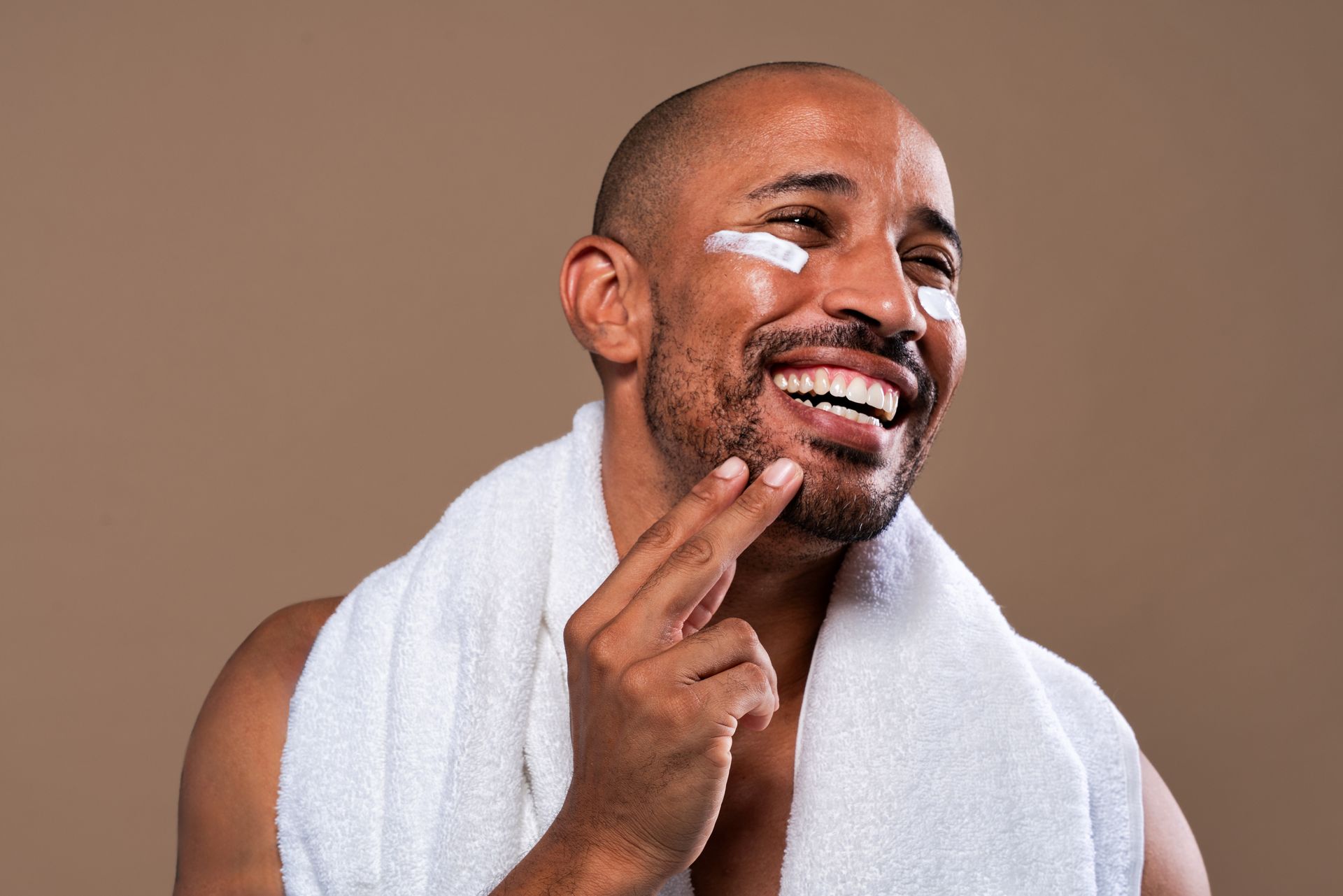 A person with a white towel around their neck smiles while applying cream to their cheek against a brown background.