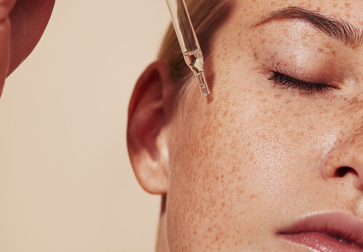 A dropper applies a clear serum to the cheek of a person with freckles against a neutral, beige background.