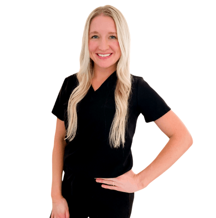 A smiling person with long blonde hair wearing black scrubs, posing with one hand on their hip against a white background.