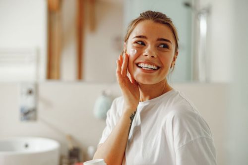 A person with a bright smile applies a dot of white cream to their cheek while standing in a bright bathroom.