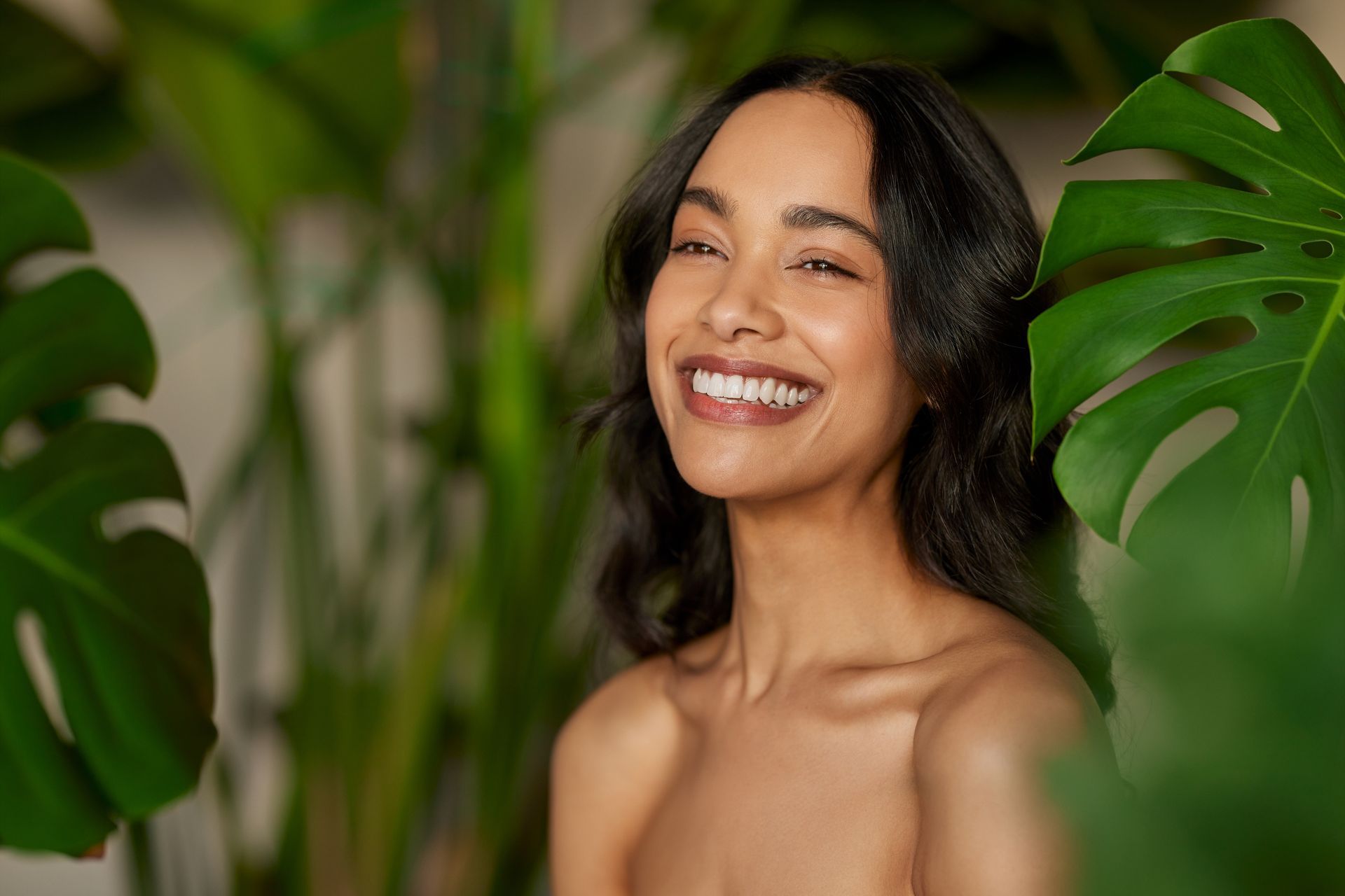 A smiling person with dark hair, framed by large green monstera leaves against a blurred botanical background.