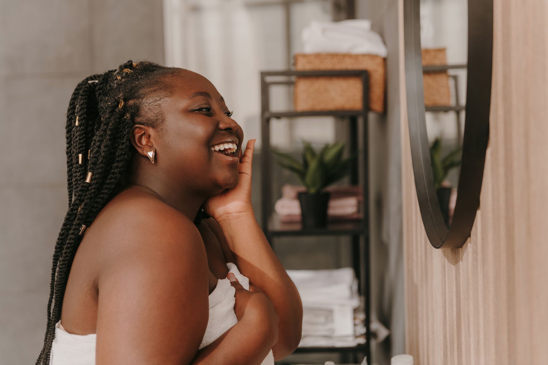 A smiling person wearing a towel wrapped around their body touches their cheek while looking into a bathroom mirror.