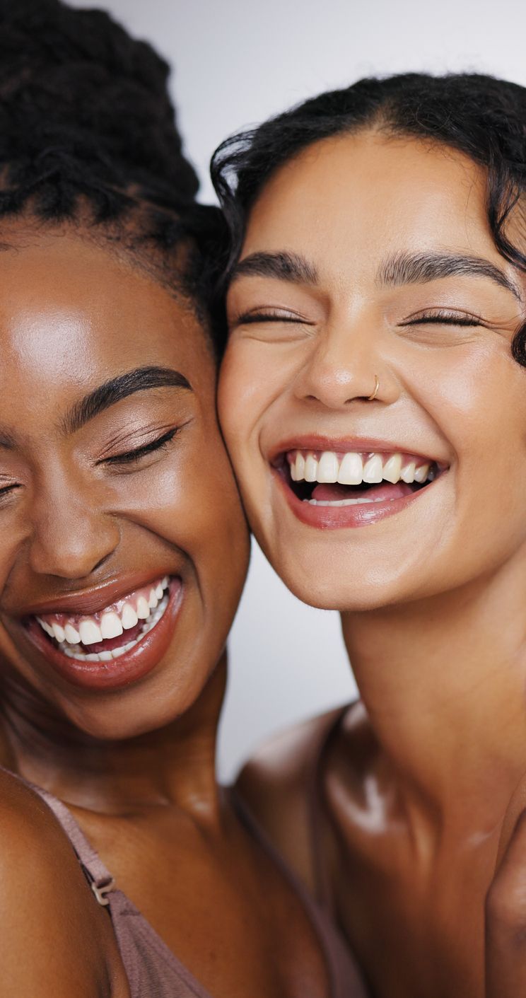 Two smiling individuals with glowing, healthy skin pose close together against a plain, light background.