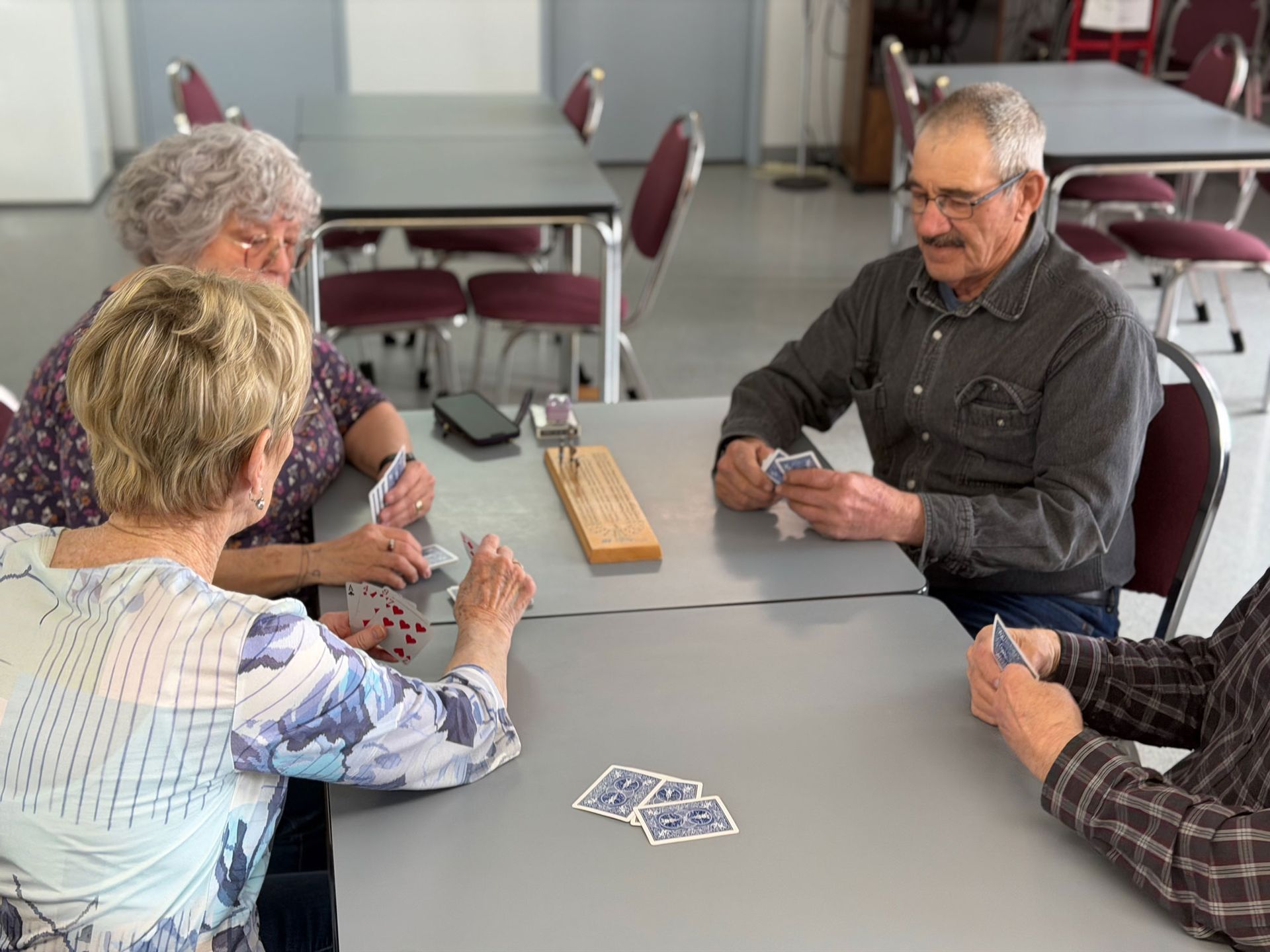 Three people sit around tables in a room playing a card game, with cards held in their hands and laid on the table.