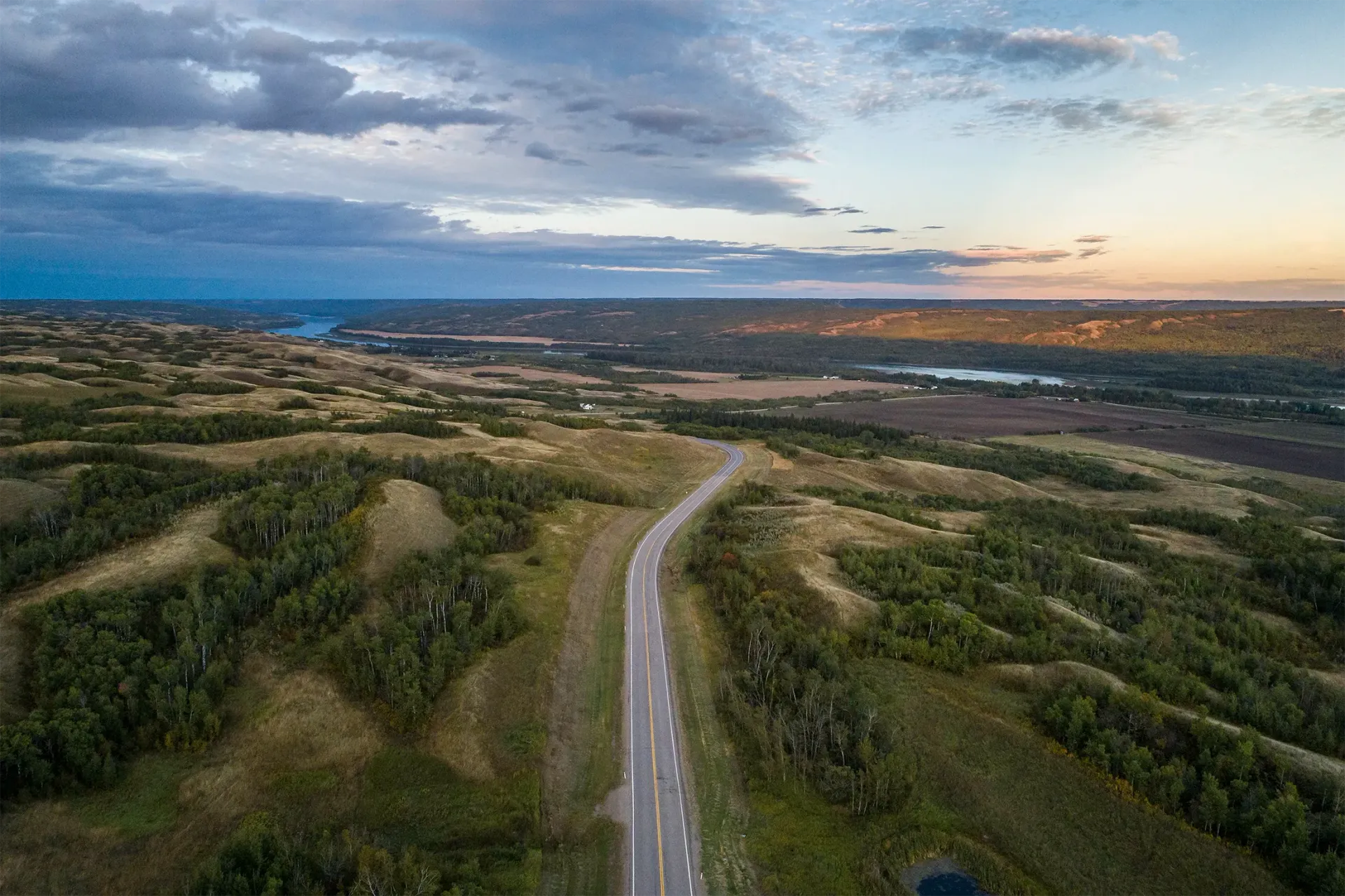 A highway stretches through a vast, rolling prairie landscape under a cloudy, sunset sky.