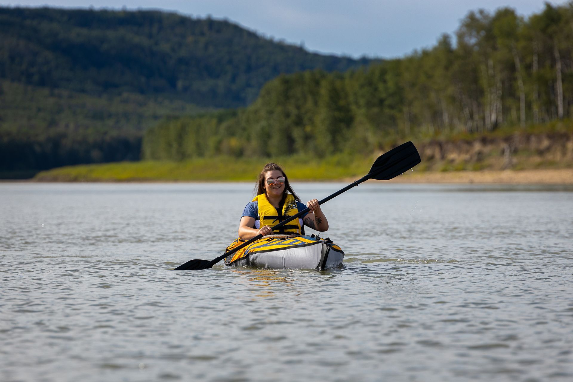A person wearing a life vest paddles a kayak on a calm river with a forested shoreline in the background.