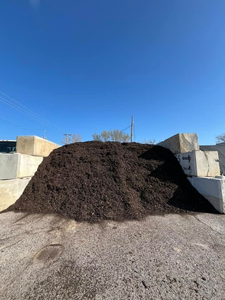 Large mound of dark mulch on a gravel lot under a clear blue sky, with concrete blocks behind it