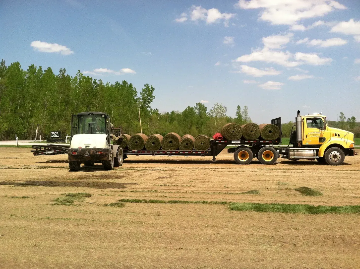 Yellow truck and white loader hauling large rolls of material across a dirt lot under a blue sky.