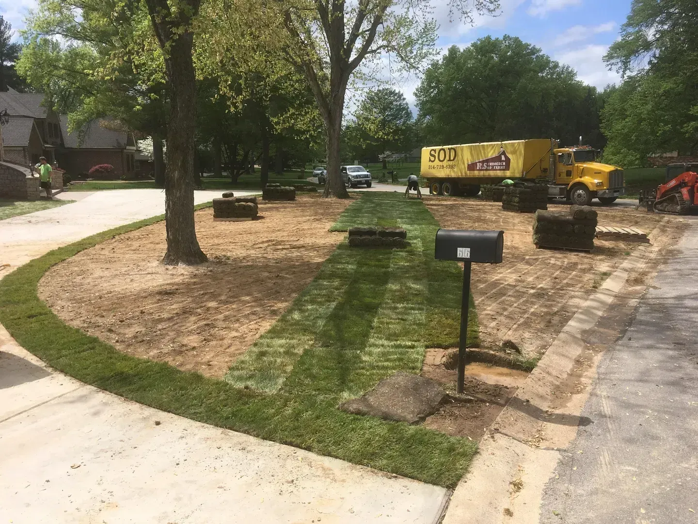 Construction equipment and mulch being installed around a landscaped yard with a mailbox and trees.