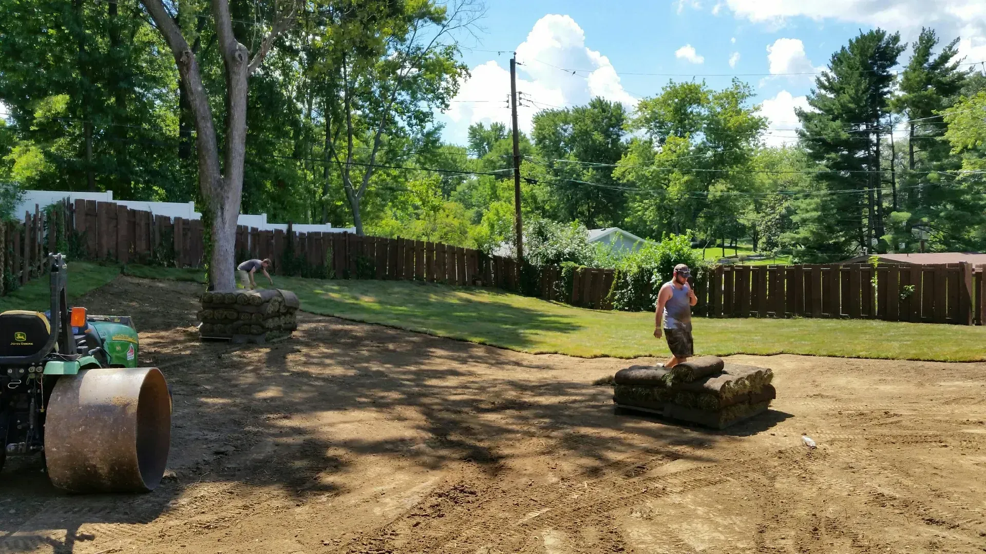 Sunny backyard with a child standing on a small dirt mound near a wooden fence and trees.