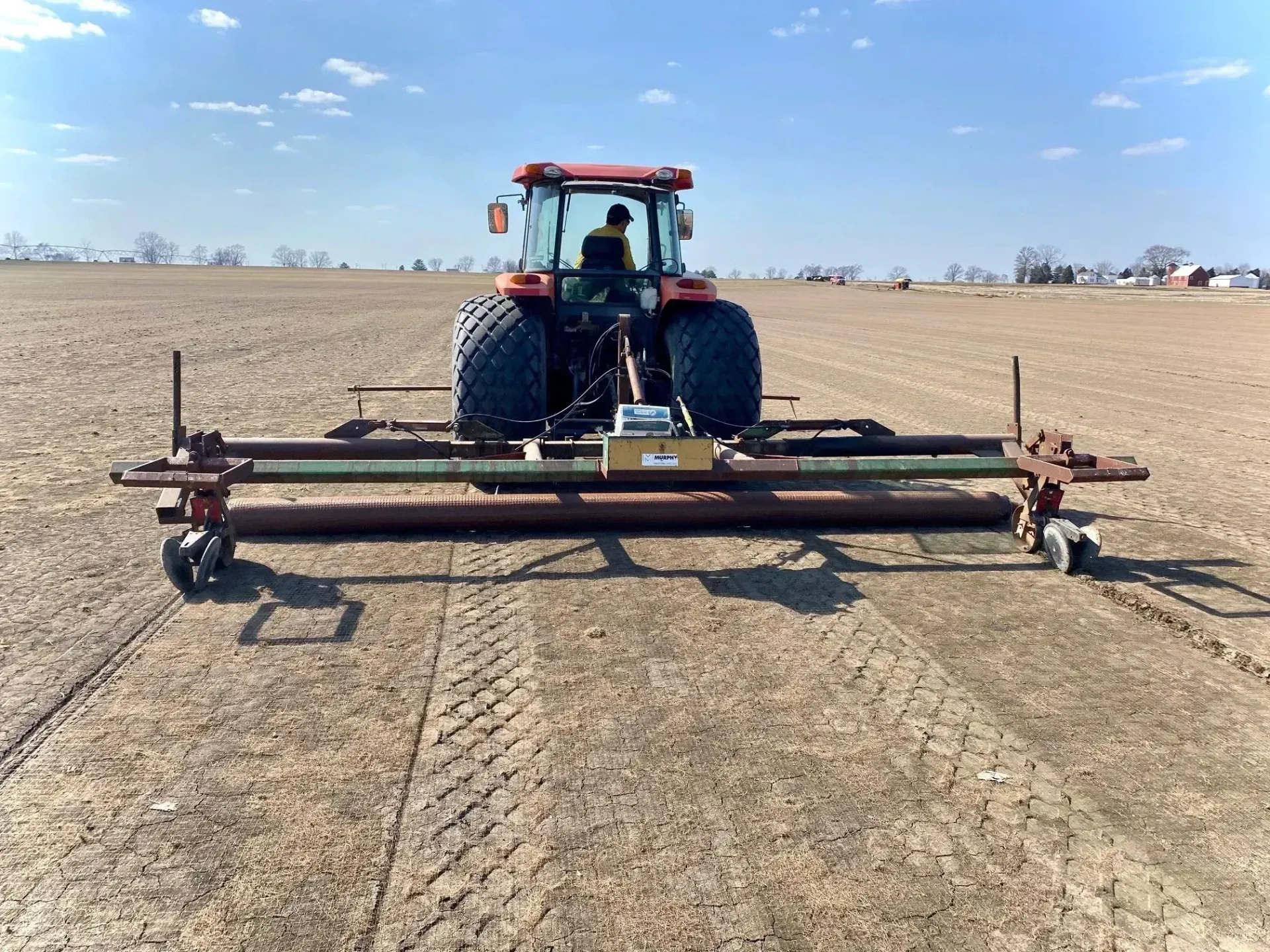 Red tractor pulling a wide farm implement across a dry, open field under a clear sky