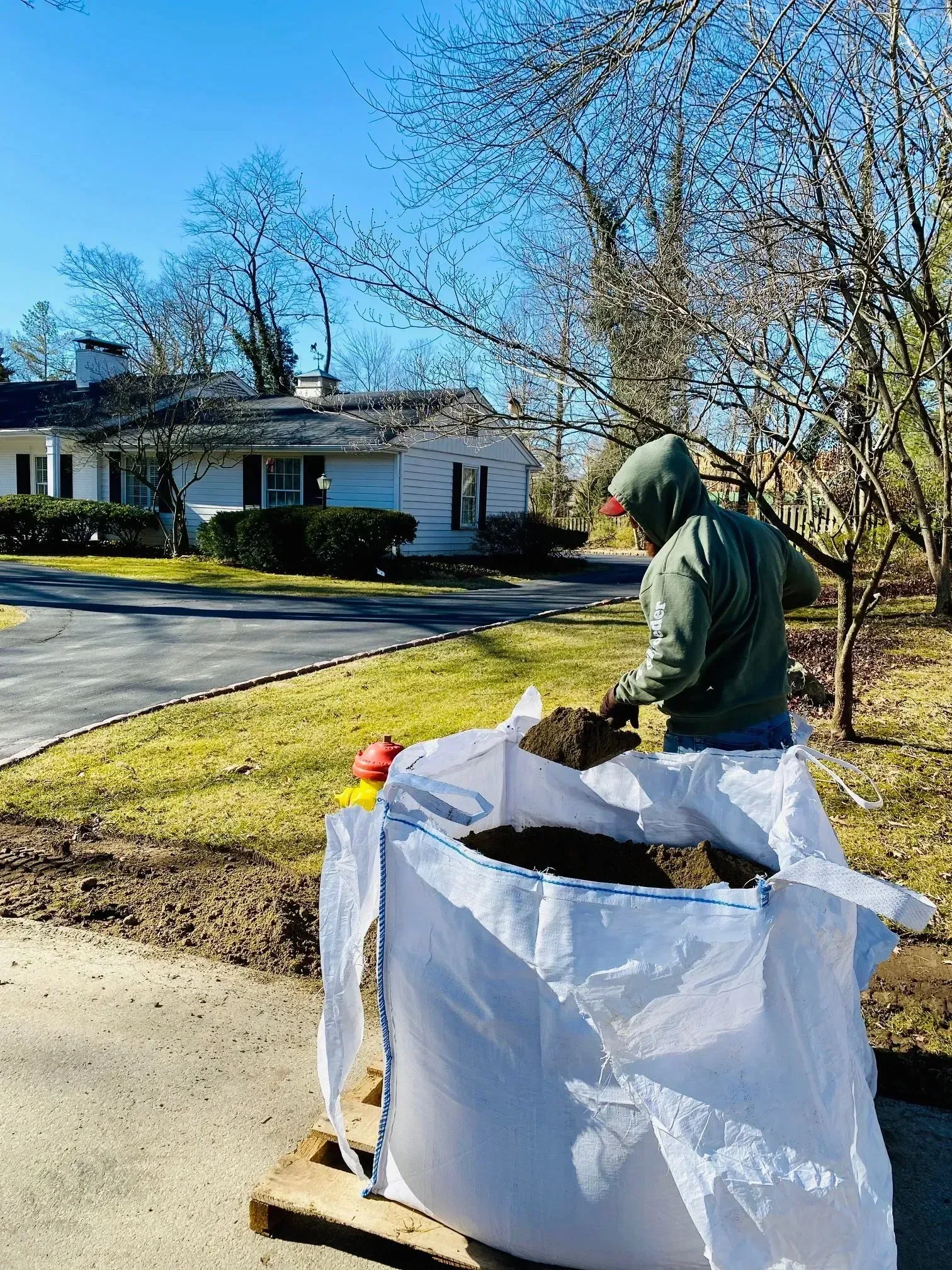 Person in a green hoodie empties yard waste into a white bag on a curb in a sunny neighborhood.