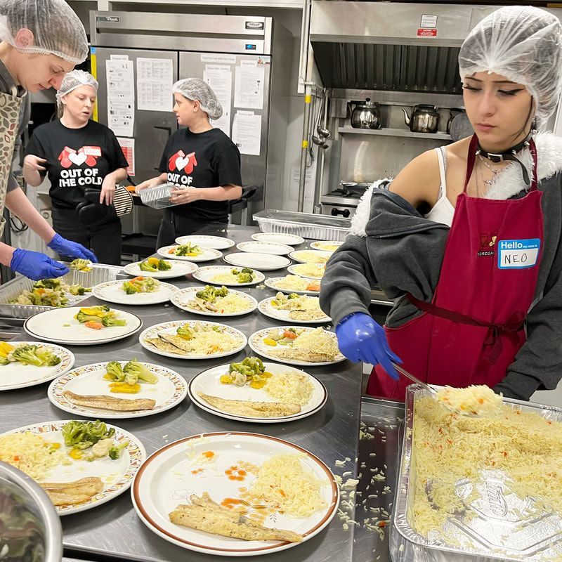 Volunteers plating meals in a commercial kitchen, with rows of prepared plates and a large tray of rice.