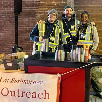 Volunteers in reflective vests smile behind an outreach table with coffee thermoses and a banner.