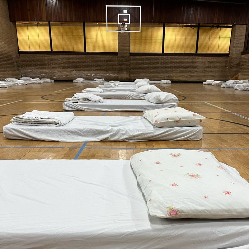 Gymnasium with rows of white sleeping mats and folded blankets spread on the floor under a basketball hoop