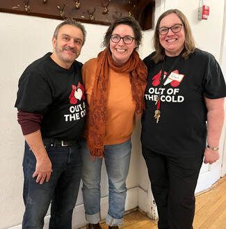 Three smiling volunteers in black T-shirts posing indoors against a white wall.
