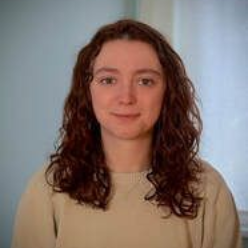 Portrait of a person with curly brown hair wearing a beige sweater against a soft blue background