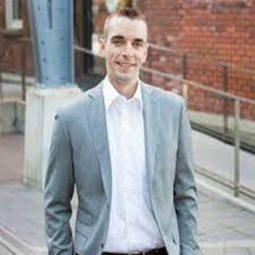 Man in a light gray blazer and white shirt standing outdoors near a brick building and railing