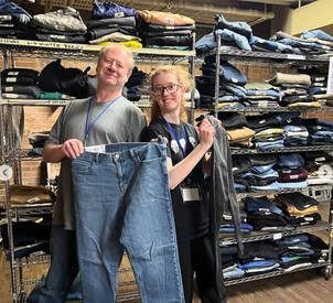 Two people smiling and holding up a pair of blue jeans in a denim shop with shelves of folded jeans