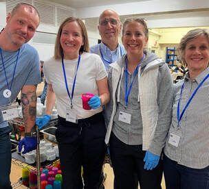 Five volunteers smiling indoors at a community event, wearing lanyards and gloves beside supplies.