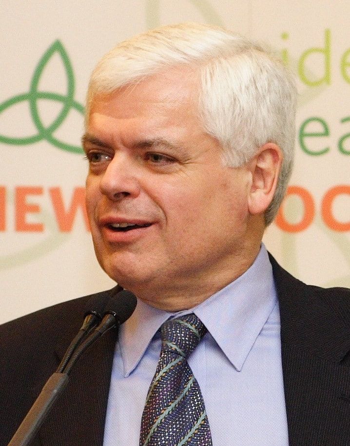 Man in a suit speaking at a podium with microphones, in front of a branded backdrop