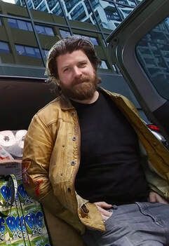 Bearded man in a tan jacket and black shirt sitting outside a modern glass building.