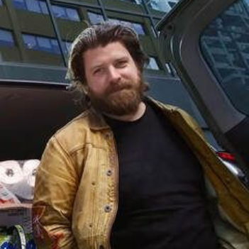 Bearded man in a tan jacket and black shirt sitting outside a modern glass building.