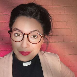 Portrait of a person wearing glasses and a clerical collar against a pink brick wall.