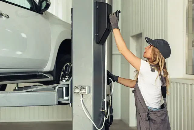 Woman in work clothes adjusting equipment near a car on a lift in a garage.