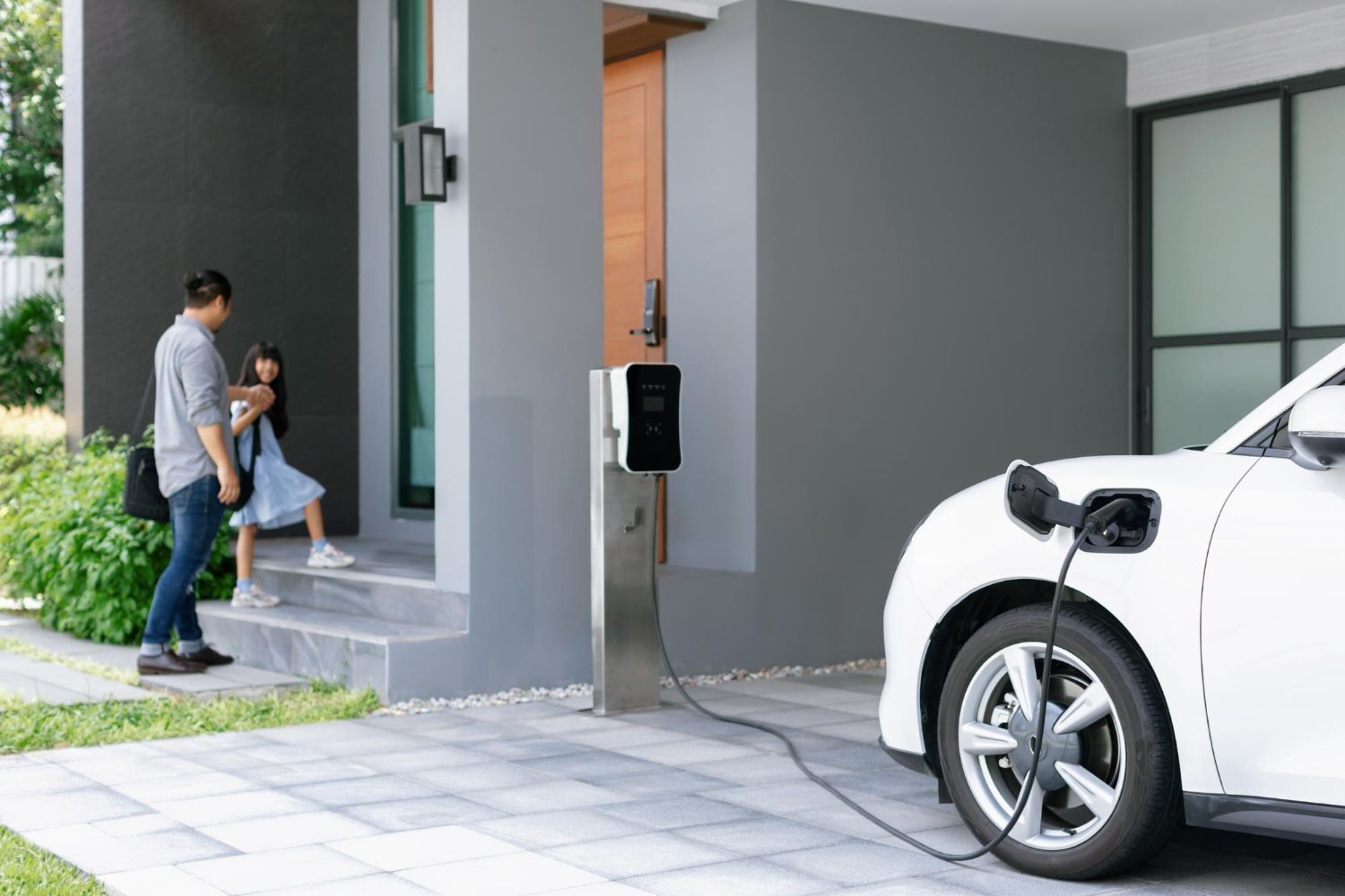 Father and child exiting a modern home. An electric car is plugged into a charger in the driveway.