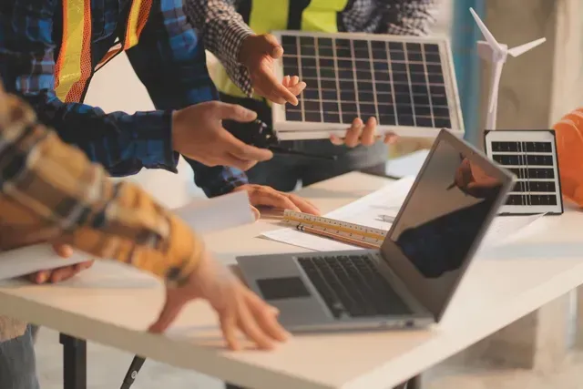 A group of people are standing around a table with a laptop and solar panels.