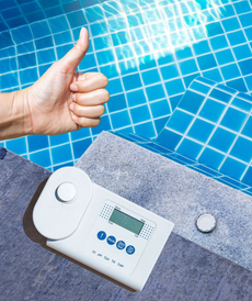 Hand giving thumbs-up beside a pool test device on the pool edge, blue tiled water in background