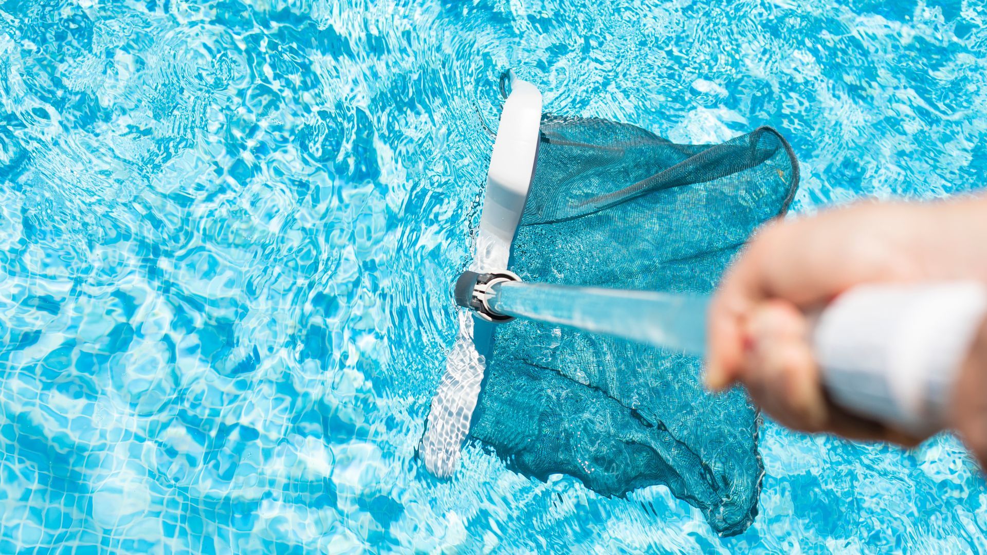 A hand holds a pool skimmer net, scooping debris from bright blue water.