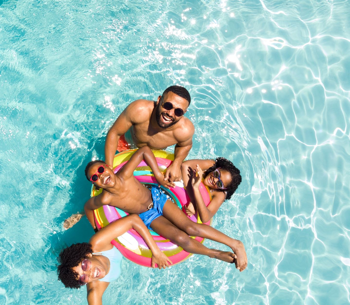 A family of four float together in a turquoise swimming pool, smiling and relaxing on colorful pool floats.