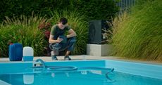 Man crouching by a backyard pool, inspecting the water near a hose and floating pool cleaner.
