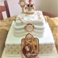 White tiered cake with gold accents and a religious topper, decorated for a celebration