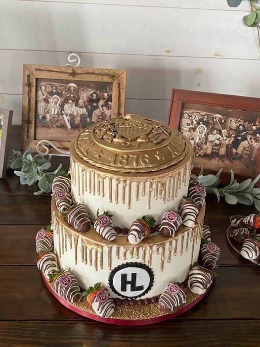 Two-tier gold-and-white cake with monogram topper and floral accents on a red board, framed photos behind