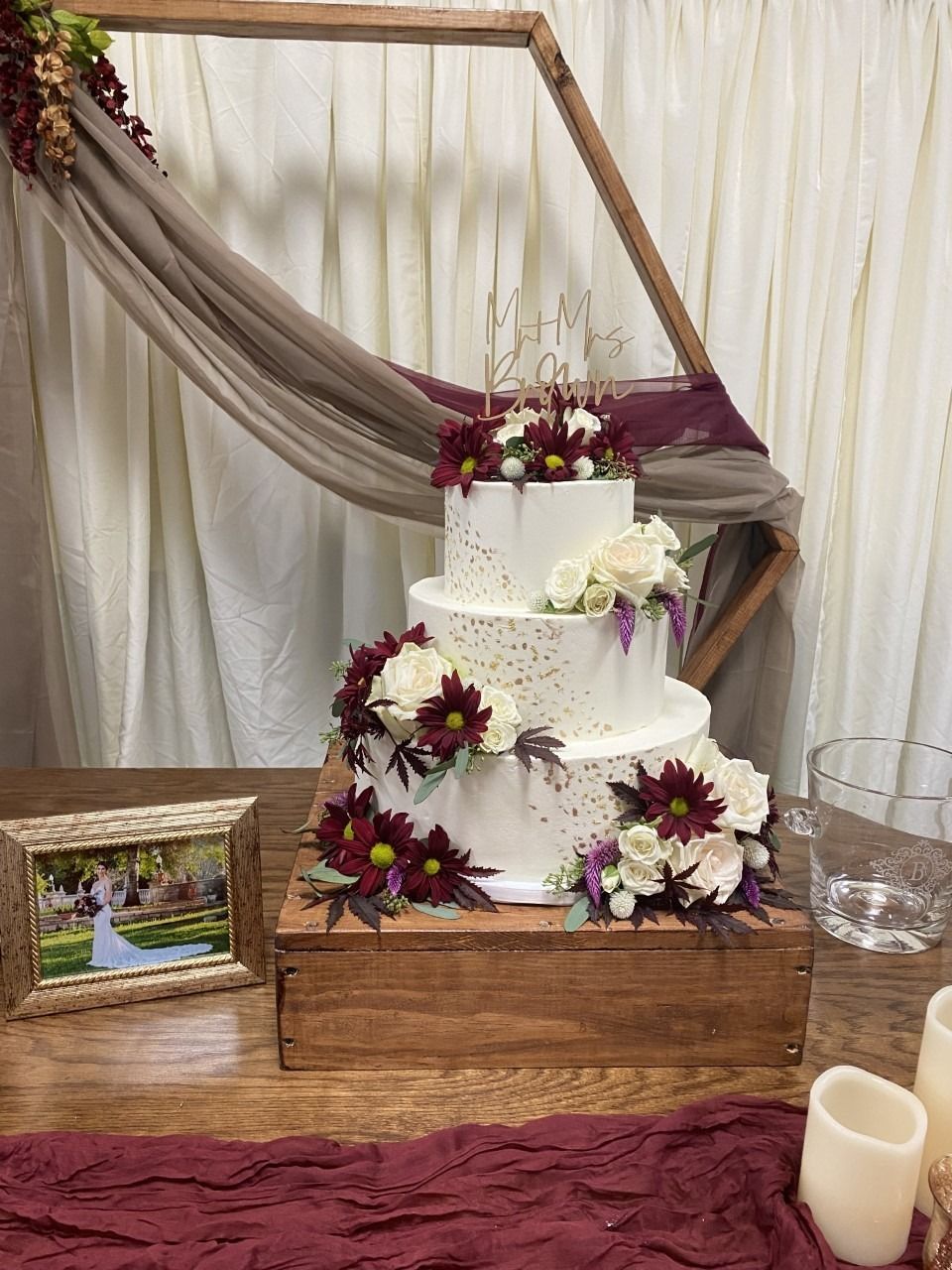 Three-tier white cake with burgundy flowers on a rustic table, draped with cream and burgundy fabric