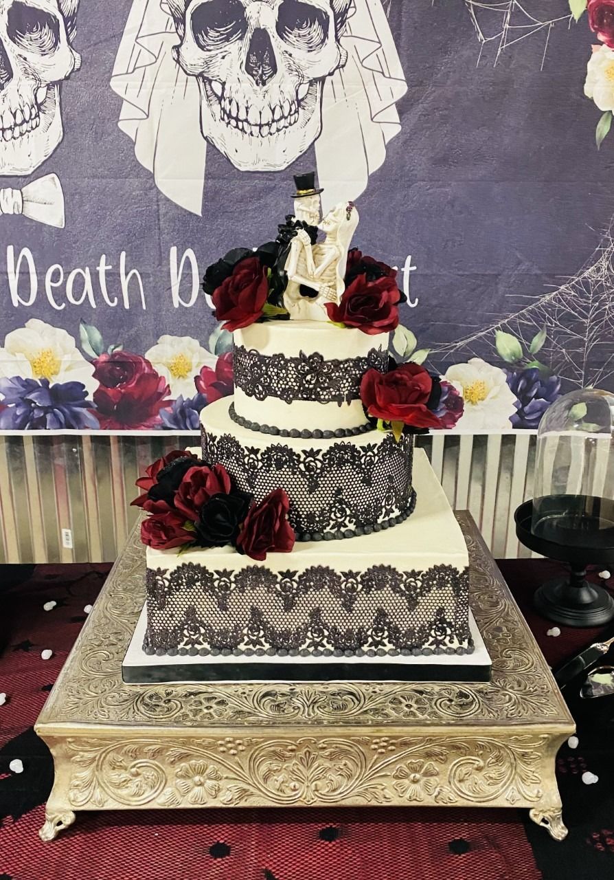 Three-tier black-and-white cake with red flowers on an ornate stand against a skull-themed backdrop