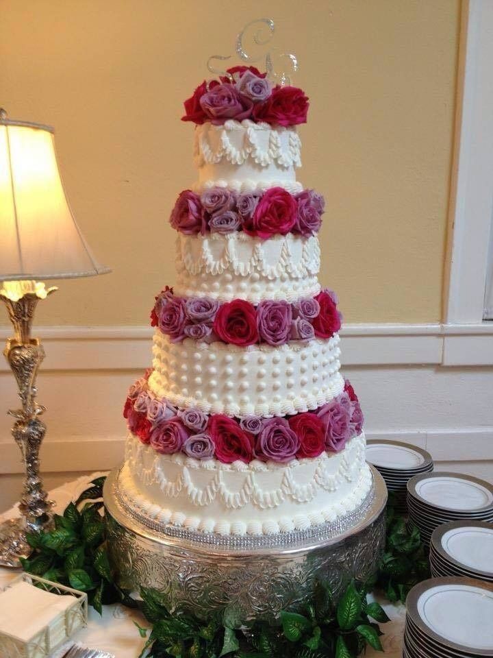 Tiered white wedding cake with pink roses on a silver stand, beside a lamp in an elegant indoor setting