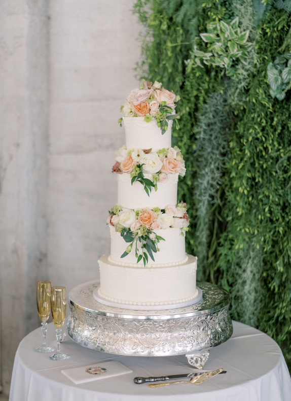 Three-tier white wedding cake with pastel flowers on a silver stand beside champagne flutes and a guest book table